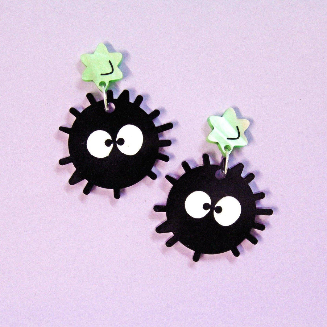 A pair of acrylic earrings with a pastel purple background. Each earring has a soot sprite (black and circular with little spines and big white eyes) hanging from a green star with faint marble patterns.