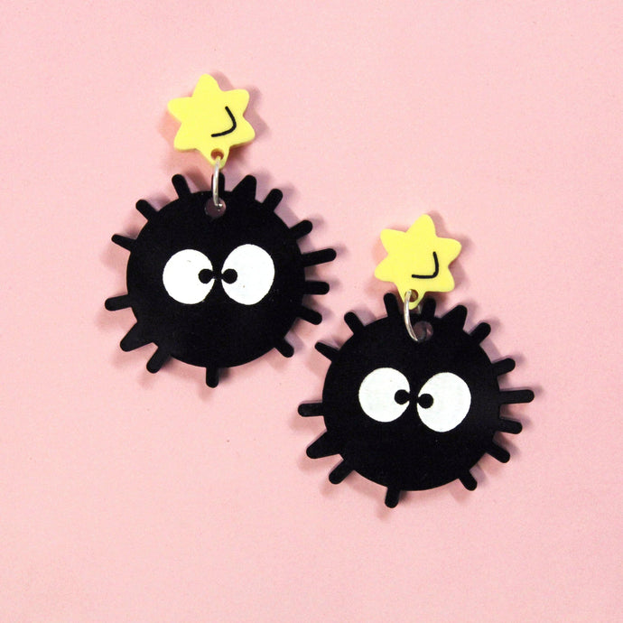 A pair of acrylic earrings in front of a rosy pink background. Each earring comprises a black ball with little hairs and big white eyes hanging from a yellow six pointed star.
