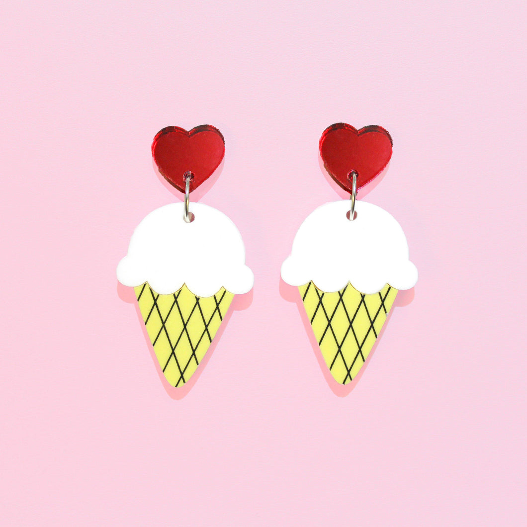 The sweetest earrings! In the shape of ice cream cones (vanilla, with pastel yellow wafer cones) hanging from two shiny red hearts like cherries on top. Ice cream earrings in front of a pastel pink background.