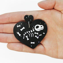 Load image into Gallery viewer, Close up of a hand holding an earring. The earring is made of two hearts: a small one attached to a large heart hanging below. Both are made from a black plastic with small, holographic glitter squares. In the middle of the large heart are scattered bones of a cartoon dinosaur with skull and claws.
