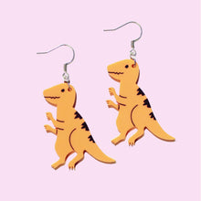 Load image into Gallery viewer, A pair of plastic orange velociraptor earrings. Each dinosaur charm hangs from a silver coloured stainless steel hook. The dinosaur is cartoonish, with narrowed eyes, sharp teeth and claws raised up, ready to pounce.
