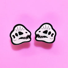 Load image into Gallery viewer, A pair of fierce dinosaur stud earrings. The dinosaur is Dilophosaurus, a carnivorous theropod that lived in the Early Jurassic with a unique crest on its head. You may also remember Dilophosaurus from Jurassic Park where he spits venom in the face of the bad guy. The earrings are in front of a pink background.
