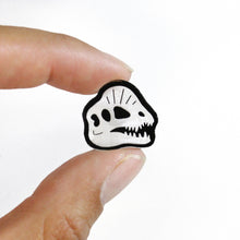Load image into Gallery viewer, A close up of the Dilophosaurus skull stud earring. The earring is small, about 2cm tall and 2cm wide and of the skull, with the bone in ghostly white and some details in black. The dinosaur is a carnivorous theropod with sharp teeth and a crest on the top of its head. The earring is held between the thumb and index finger by a woman in front of a white background.

