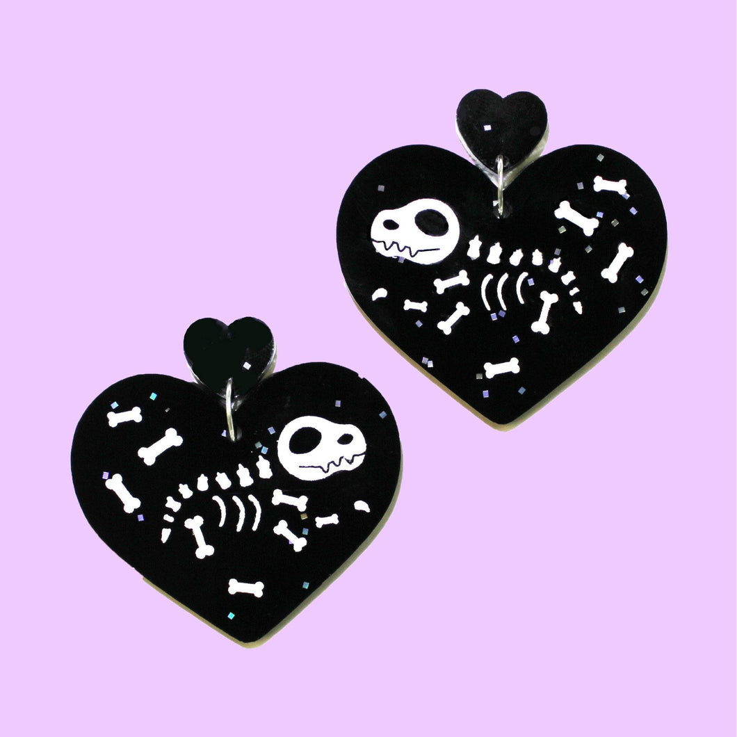 A pair of dangly earrings with dinosaur skeletons. The earrings are mirror images and made of two parts: a small heart charm with a large heart bearing dinosaur skeleton in white hanging below. Both hearts are made from acrylic with speckled squares of glitter.
