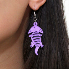 Load image into Gallery viewer, A close up of an earring of Diplocaulus, an extinct amphibian that looks like a salamander with a boomerang shaped head. The earring is made from acrylic, a type of plastic, that is light purple with silver glitter and hand painted details including black stripes along its back. The earring is hanging from a silver-coloured stainless steel hook and is worn by a biracial woman with long, dark brown hair.

