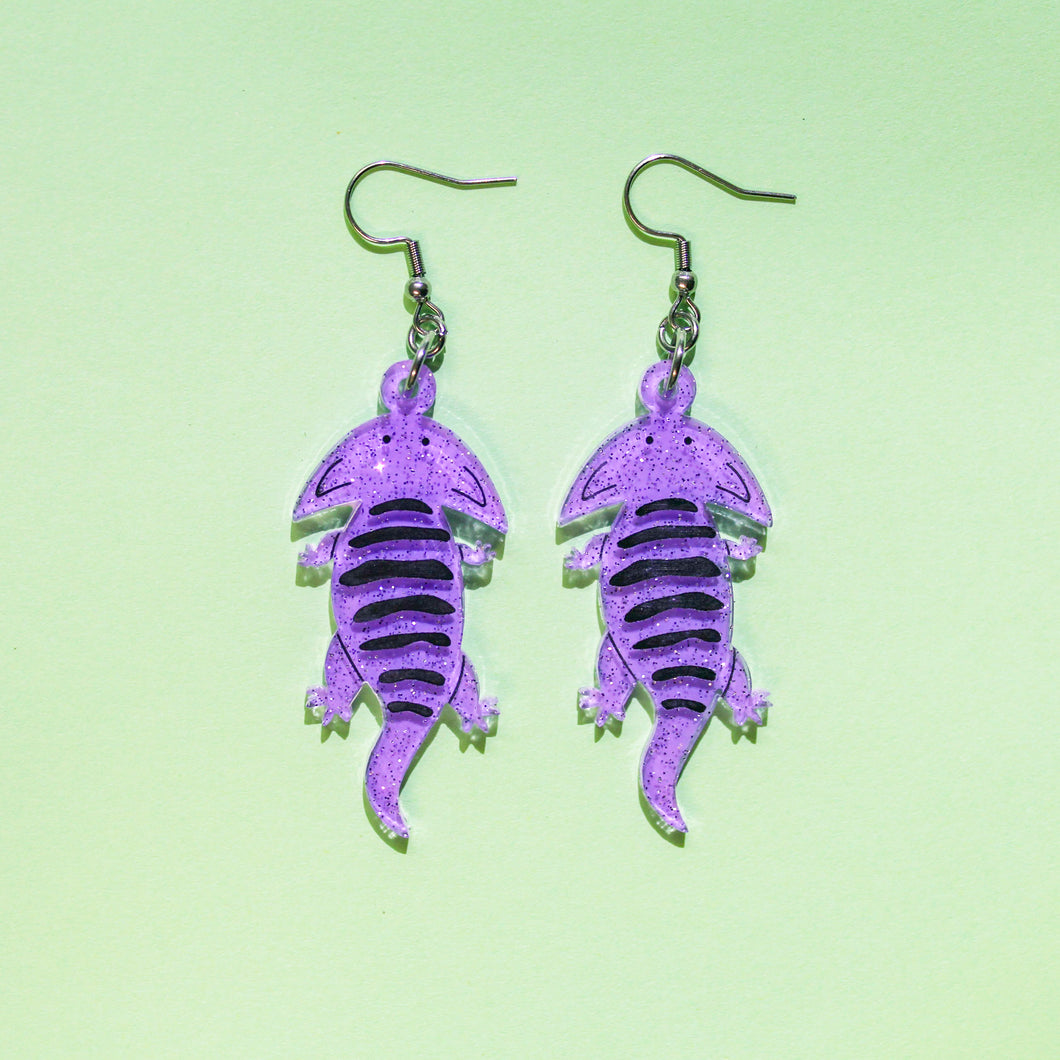 A pair of acrylic earrings of Diplocaulus, an extinct amphibian that lived long before the dinosaurs during the Permian in what is now North America and Africa. Diplocaulus looks like a salamander with a boomerang shaped head. The earrings are made from acrylic, a type of plastic and are light purple with glittery sparkles. There is some hand painted details, including the eyes and black stripes along the back.