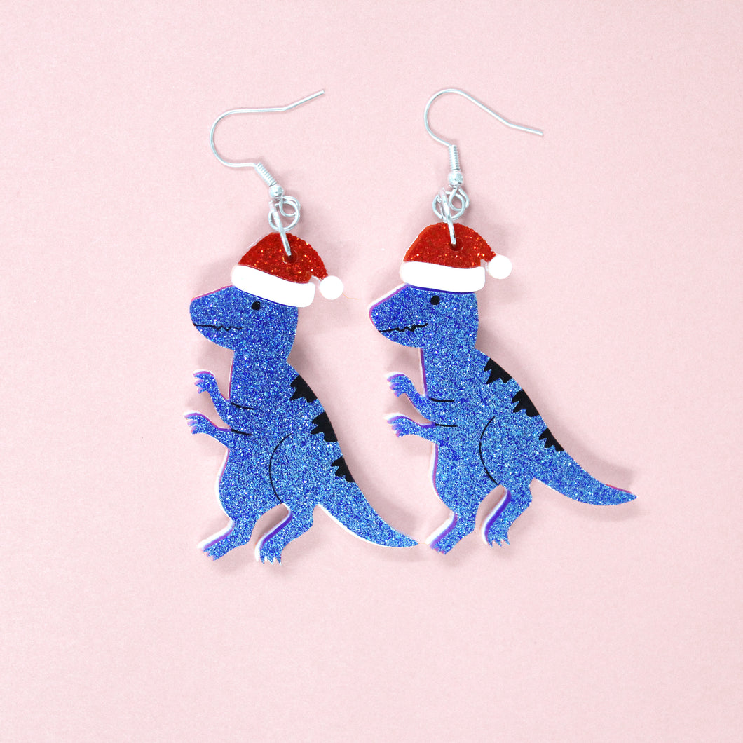 A pair of Raptor dinosaur earrings. The raptors are cartoons, with narrowed eyes, jagged teeth and little 3 fingered hands. The raptors are a pair in glittery blue and wearing sparkly red Santa hats, ready for Christmas. They hang from silver coloured hypoallergenic hooks in front of a baby pink background.
