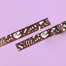 Load image into Gallery viewer, Sections of washi tape, which features dinosaur bones in white, in front of a slightly ombre brown background and surrounded by holographic foil sparkles. The washi tape is 5mm wide, and each roll is 10m thick. Good for decorating planners and diaries. The tape is in front of a pastel purple background.
