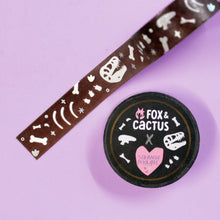 Load image into Gallery viewer, A roll of washi tape, which features dinosaur bones in white, in front of a slightly ombre brown background and surrounded by holographic foil sparkles. The washi tape is 5mm wide, and each roll is 10m thick with a special sticker that says &quot;Fox &amp; Cactus X Strange Magic&quot;. The tape is in front of a pastel purple background.

