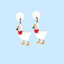 Load image into Gallery viewer, A pair of earrings featuring white geese proudly wearing a red bow around the neck and holding a shiny gold coloured bell. The earrings are made from acrylic, a type of plastic and are in front of a baby blue background.
