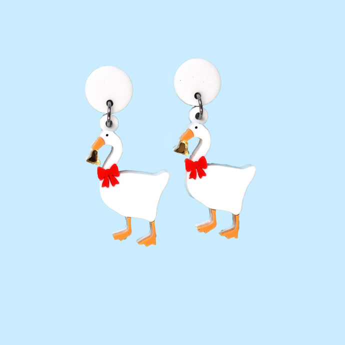 A pair of earrings featuring white geese proudly wearing a red bow around the neck and holding a shiny gold coloured bell. The earrings are made from acrylic, a type of plastic and are in front of a baby blue background.