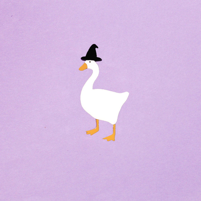 A cute lil brooch of a goose wearing a tiny black witch hat. Made from acrylic plastic, in front of a purple background. The goose is plain white, with an orange beak and matching orange legs.