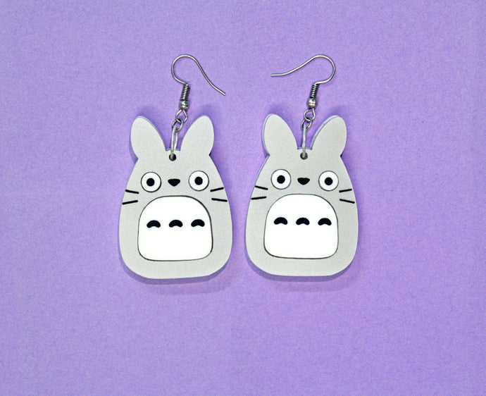 A pair of earrings in front of a purple background. The earrings feature a large plastic charm of an owl, rabbit, cat hybrid hanging from silver coloured hooks.