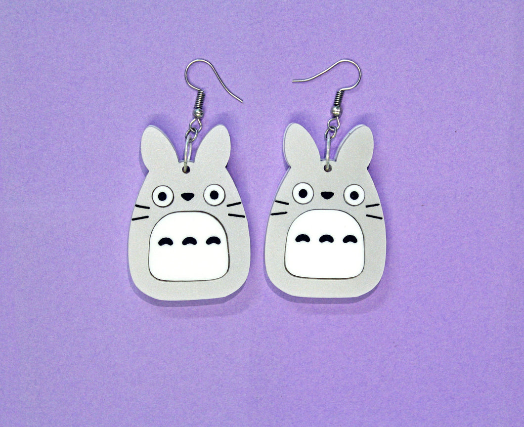 A pair of earrings in front of a purple background. The earrings feature a large plastic charm of an owl, rabbit, cat hybrid hanging from silver coloured hooks.