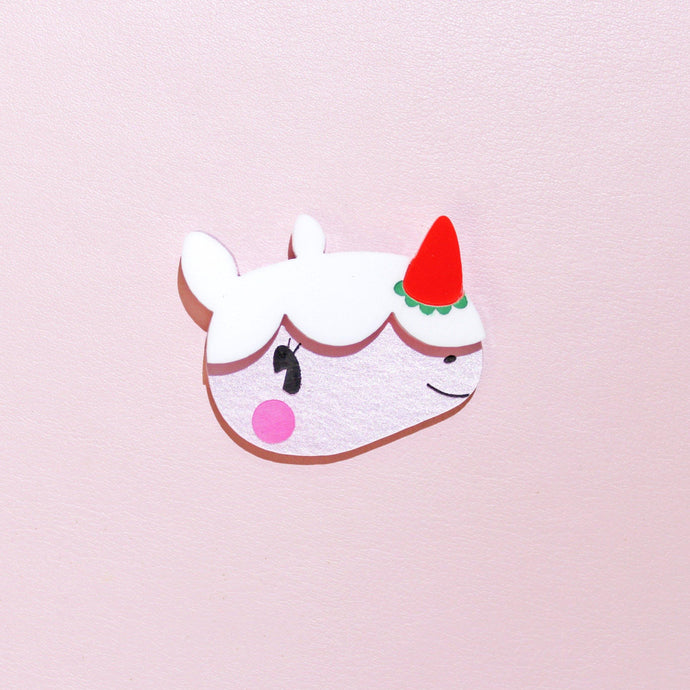 A close up of a unique looking brooch. The brooch is made from acrylic plastic and is of a cute, cartoon rhino. She has pink blushing cheeks, a strawberry for a horn and whipped icing on top of her head with a little smile on her face. Her name is Merengue, and is in front of a pale pink background.
