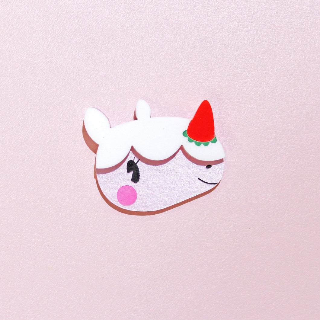 A close up of a unique looking brooch. The brooch is made from acrylic plastic and is of a cute, cartoon rhino. She has pink blushing cheeks, a strawberry for a horn and whipped icing on top of her head with a little smile on her face. Her name is Merengue, and is in front of a pale pink background.