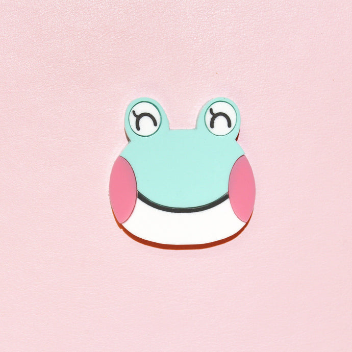 A close up of a cute frog brooch. The frog's name is Lily, and she has her eyes closed, large pink cheeks, a mint green head and white chin separated by a thin mouth. She's made from acrylic, a type of plastic and is in front of a millenial pink background.