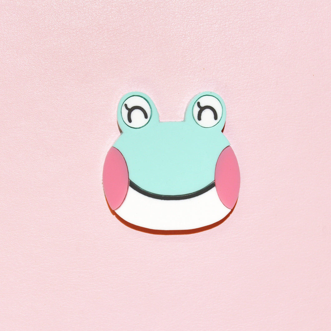 A close up of a cute frog brooch. The frog's name is Lily, and she has her eyes closed, large pink cheeks, a mint green head and white chin separated by a thin mouth. She's made from acrylic, a type of plastic and is in front of a millenial pink background.