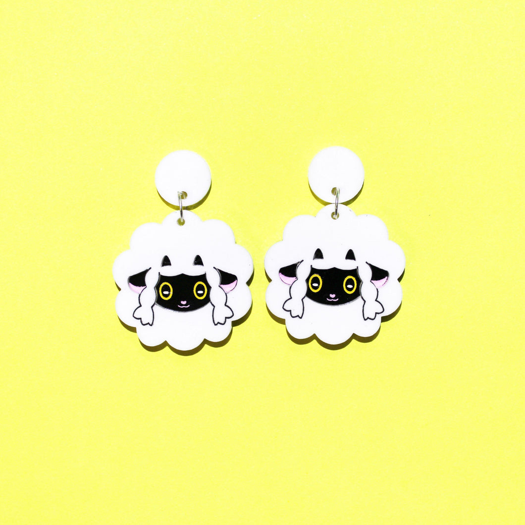 A pair of earrings of cartoon sheep with white wool, little black horns, braids either side of their black faces and yellow eyes. The sheep earrings are in front of a yellow background.