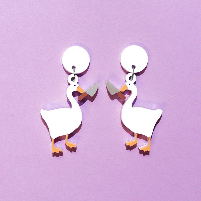 A pair of plastic earrings featuring geese holding large knives. The geese are white and mirror images, with orange bills and webbed feet on a purple background.