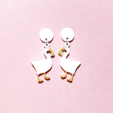 Load image into Gallery viewer, A pair of earrings featuring white geese holding shiny gold coloured bells. The earrings are in the middle of the picture with a pastel pink background.
