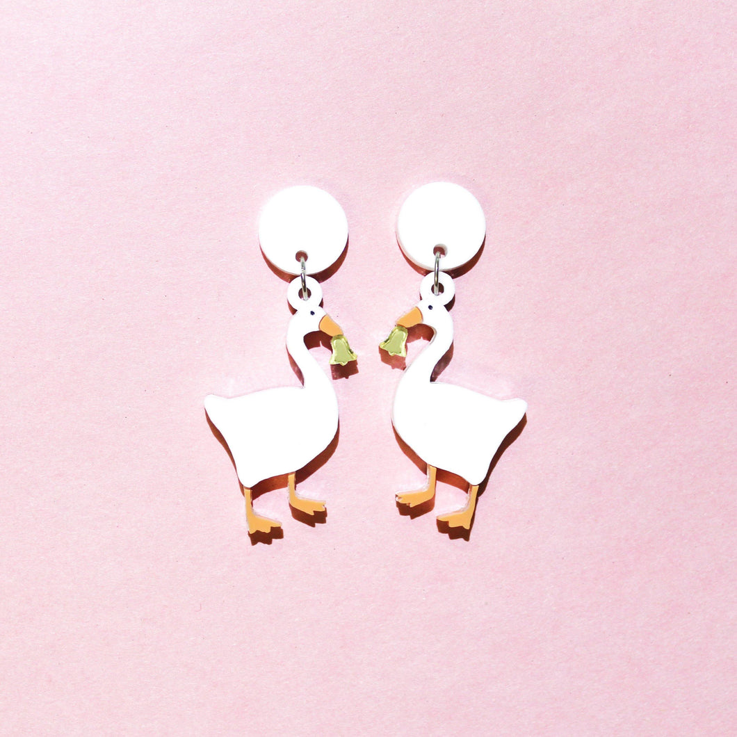 A pair of earrings featuring white geese holding shiny gold coloured bells. The earrings are in the middle of the picture with a pastel pink background.