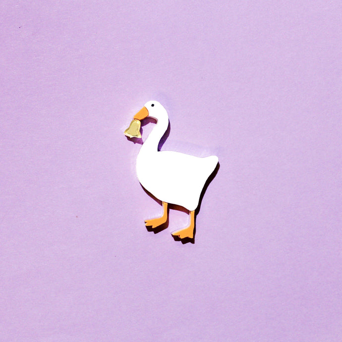 Plastic brooch of a white goose holding a gold coloured bell in its beak on a purple background