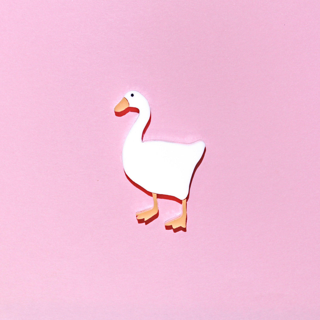 A plastic brooch of a white goose with orange bill and webbed feet on a pink backdrop.