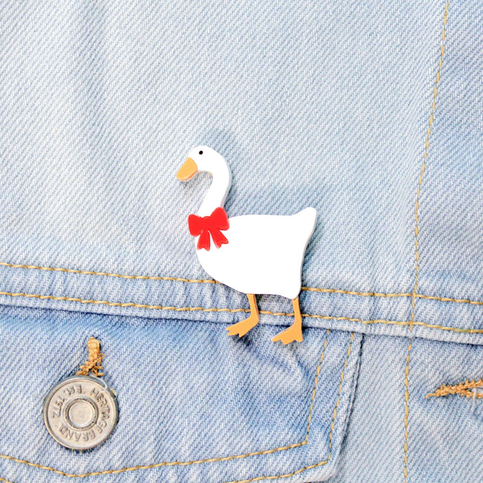 Close up of a brooch on a pale blue denim jacket. The brooch is made of a type of plastic, featuring a white goose with orange feed and beak with a red bow around its neck.