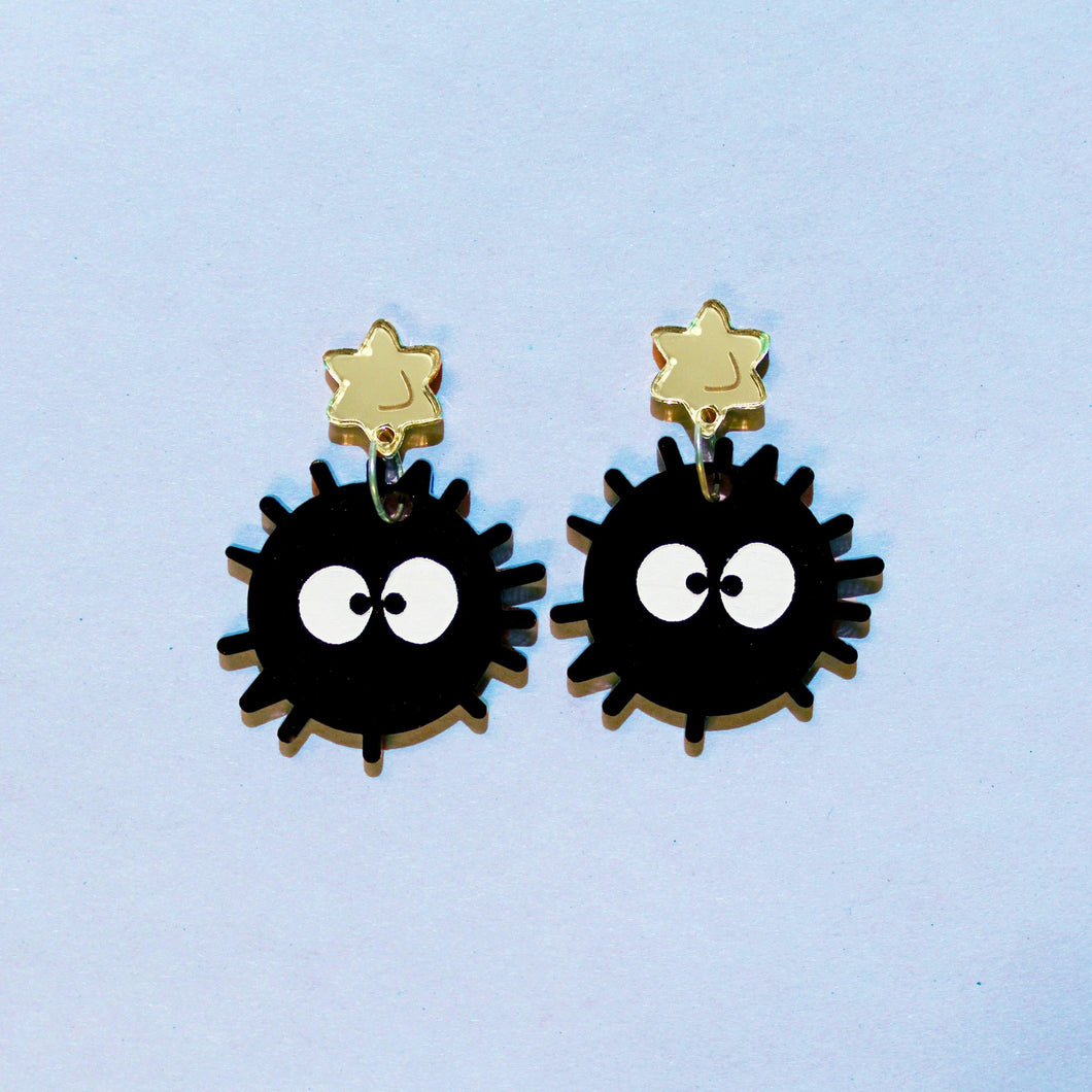 A pair of earrings in front of a blue background. They are based on imaginary sprites or dust balls, susuwatari hanging from plastic gold coloured stars. The soot sprites are black balls with spine-like projections and big white eyes.