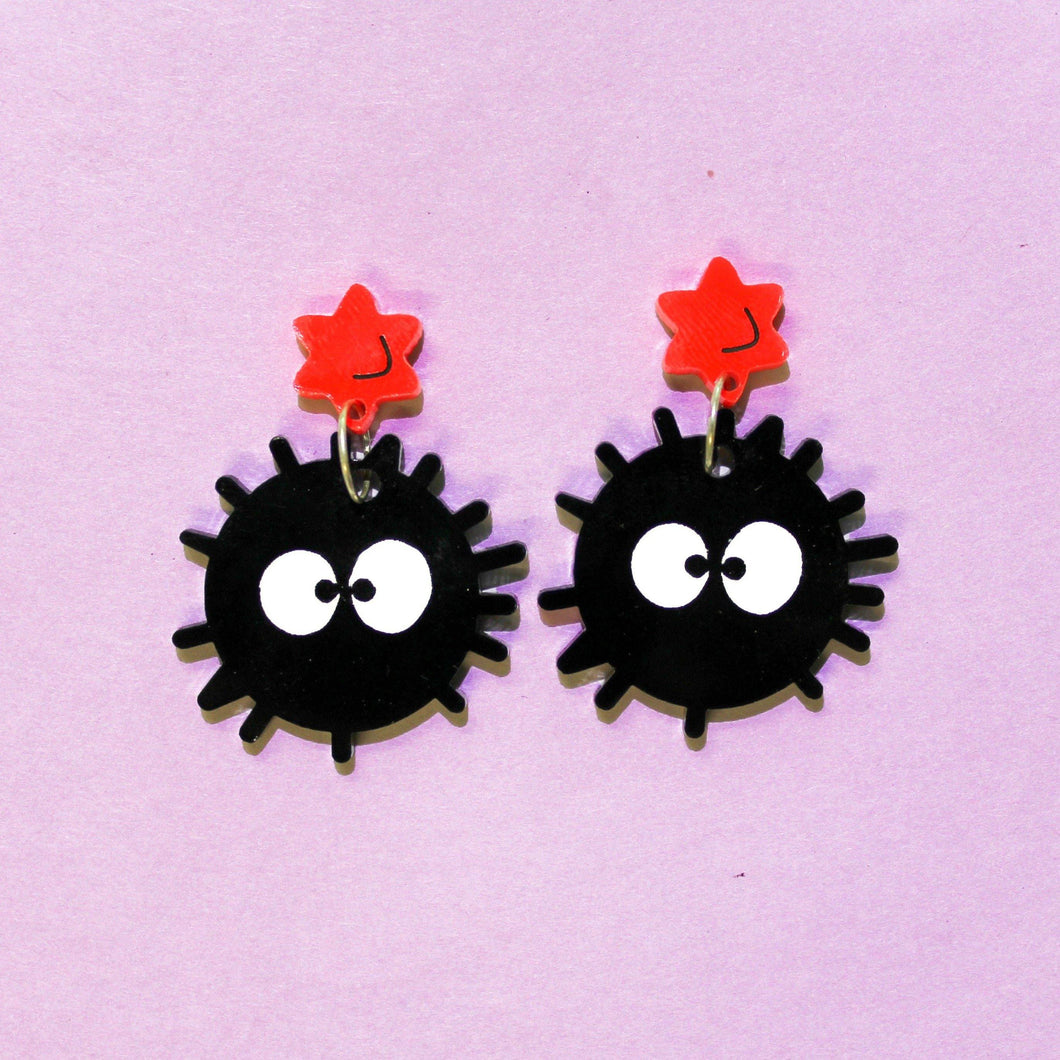 A pair of acrylic plastic earrings of soot sprites; imaginary dust balls with white googly eyes hanging from bright red stars from silver rings. Behind them is a purple background.