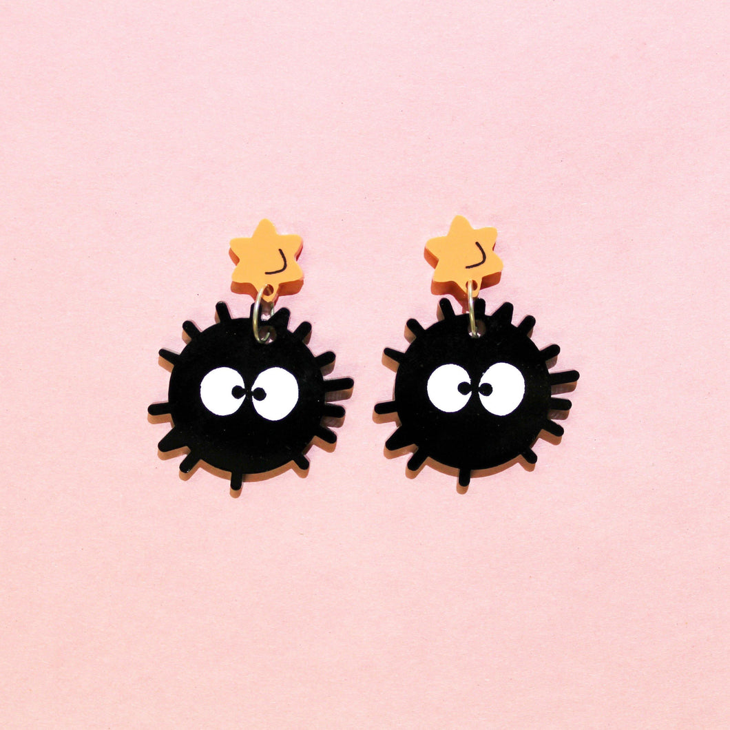 A pair of soot sprite earrings; fictitious creatures resembling black balls with big white eyes and little hairs. They hang from orange stars in front of a pink background.