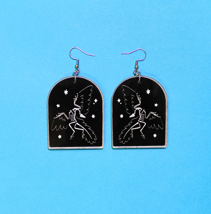 Tombstone shaped earrings on a blue background. The arch or tombstone shape is black with a faint white border and the dinosaur Archaeopteryx also in white, surrounded by stars. Archaeopteryx is the famous feathered dinosaur, and in this depiction, its head is thrown back dramatically in the 'death pose'. The earrings are made from acrylic, a type of plastic with silver-coloured stainless steel hooks.