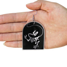 Load image into Gallery viewer, A close up of a dinosaur earring in a hand. The earring is a skeleton of the dinosaur Tyrannosaurus rex made from acrylic, a type of plastic. The charm is black, and the bones of the animal are in a transparent white. The earring is in the hand of a woman, in front of a white background.
