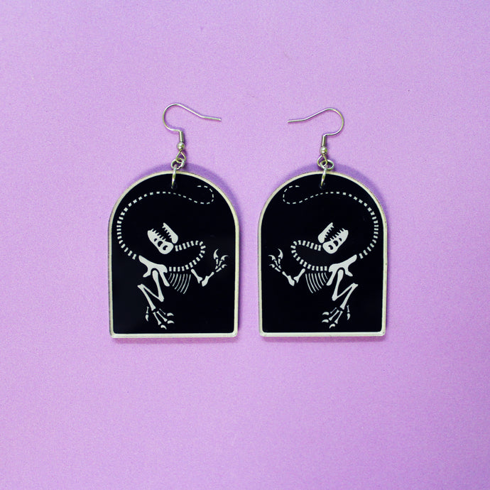 Tombstone shaped earrings on a purple background. The arch or tombstone shape is black with a faint white border and the dinosaur T rex also in white. In this depiction, T rex has its head thrown back dramatically in the 'death pose'. The earrings are made from acrylic, a type of plastic with silver-coloured stainless steel hooks.