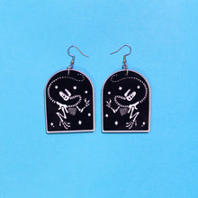 Load image into Gallery viewer, Tombstone shaped earrings on a blue background. The arch or tombstone shape is black with a faint white border and the dinosaur T rex surrounded by stars also in white. In this depiction, T rex has its head thrown back dramatically in the &#39;death pose&#39;. The earrings are made from acrylic, a type of plastic with silver-coloured stainless steel hooks.
