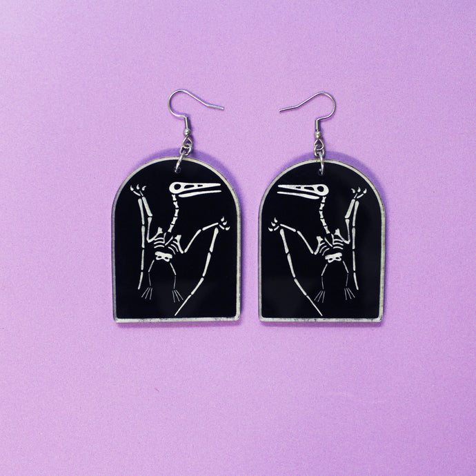 Tombstone shaped earrings on a purple background. The arch or tombstone shape is black with a faint white border and a pterodactyl also in white. The earrings are made from acrylic, a type of plastic with silver-coloured stainless steel hooks.