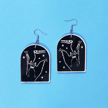 Load image into Gallery viewer, Tombstone shaped earrings on a blue background. The arch or tombstone shape is black with a faint white border and a pterodactyl surrounded by stars also in white. The earrings are made from acrylic, a type of plastic with silver-coloured stainless steel hooks.

