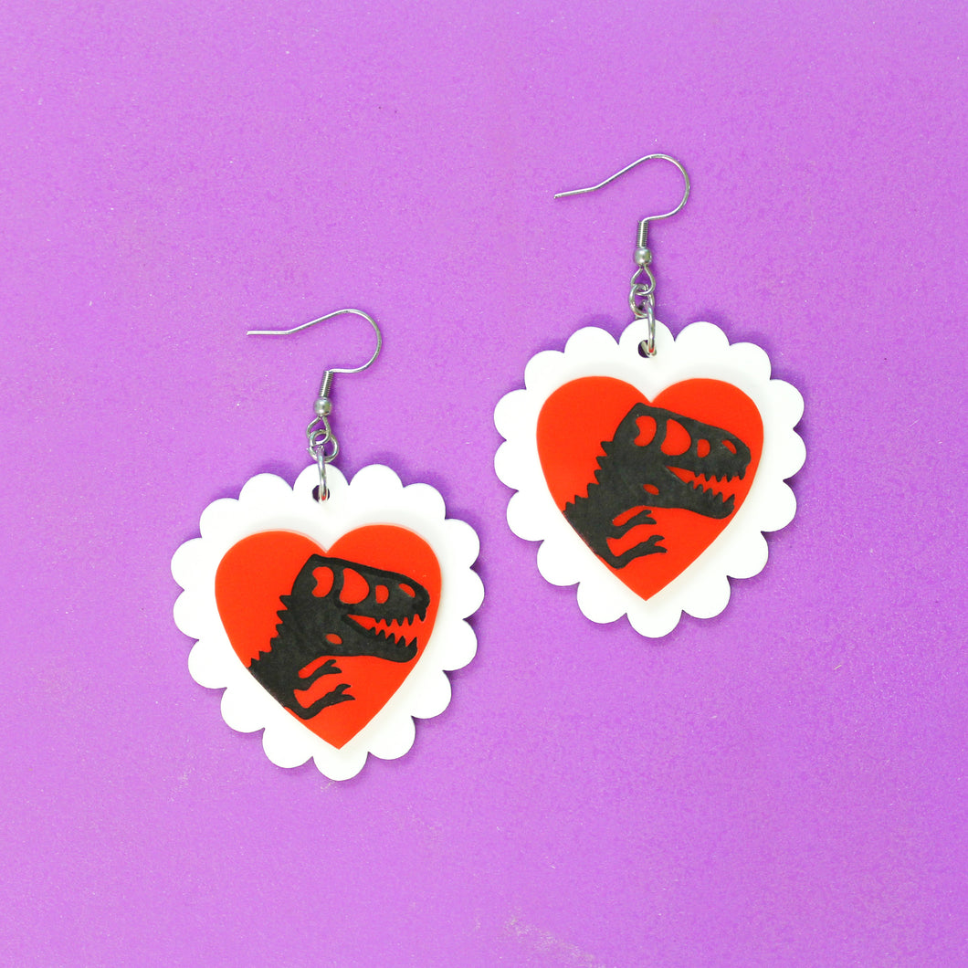 A pair of acrylic (plastic) earrings, featuring a dinosaur silbouette on a red heart, surrounded by white scalloped edges. Inspired by a famous dinosaur movie and vintage valentine's day cards. The dinosaur silhouette is handpainted. The earrings hang from stainless steel hooks.