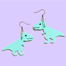 Load image into Gallery viewer, A pair of mint T Rex earrings. The T Rex are cartoonish with jagged closed mouths, narrow eyes and small front arms with two claws on each hand. The T Rex hang from silver coloured earring hooks in front of a light purple background.
