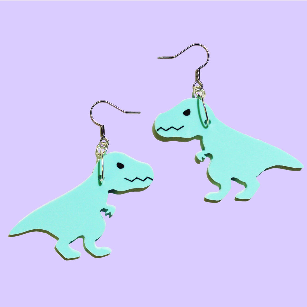 A pair of mint T Rex earrings. The T Rex are cartoonish with jagged closed mouths, narrow eyes and small front arms with two claws on each hand. The T Rex hang from silver coloured earring hooks in front of a light purple background.