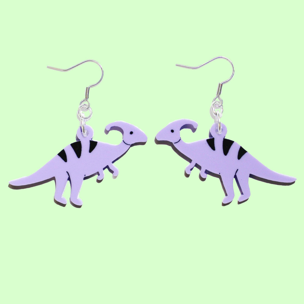 A pair of light purple coloured dinosaur earrings. The earrings are Parasaurolophus, a herbivore standing up on two legs with a bizarre head that looks a bit like a comma. Or just think of Ducky, from the Land Before Time. The earrings are made from acrylic and hang from stainless steel hooks in front of a light green background.