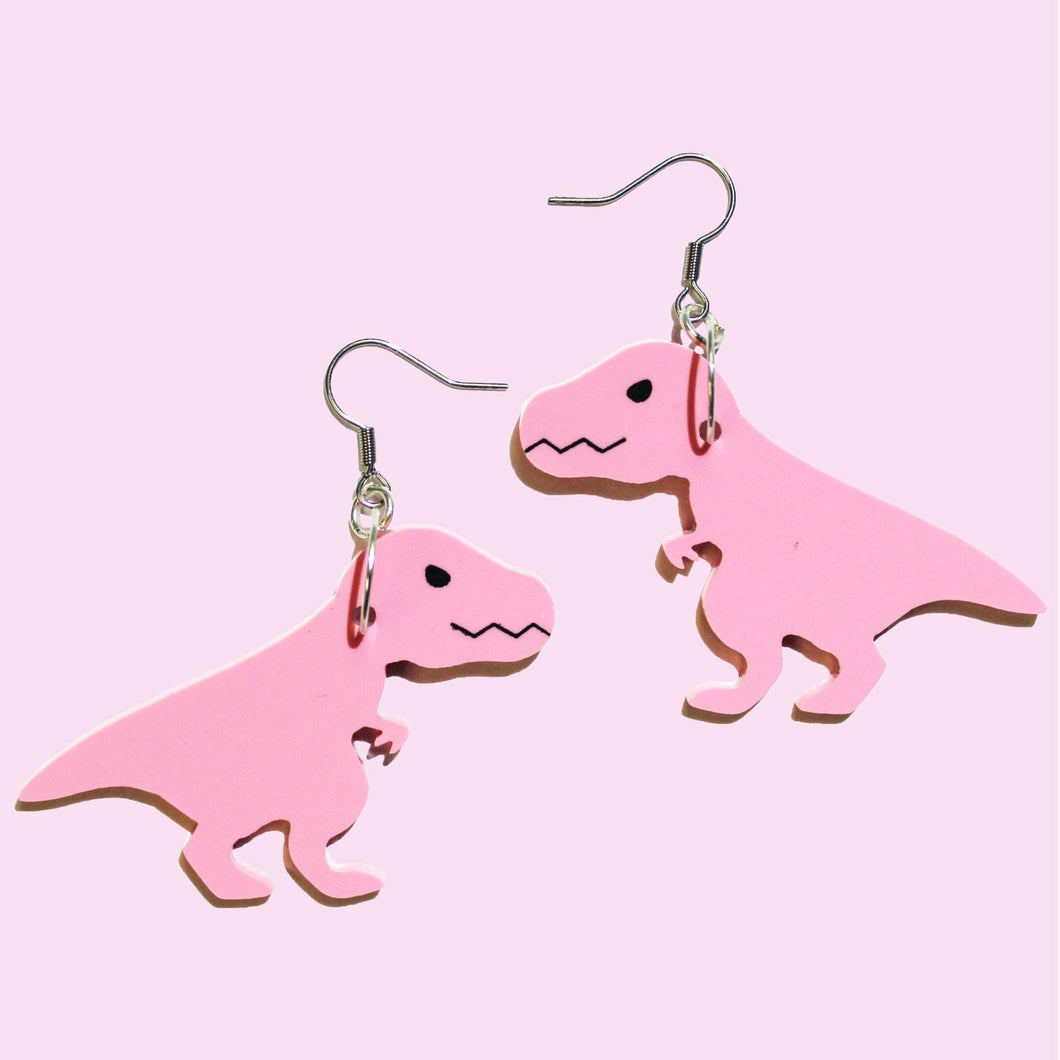 A pair of cartoon, pink T rex earrings in front of a pastel pink background. The earrings are mirror images of one another, both hanging from silver coloured stainless steel hooks. 