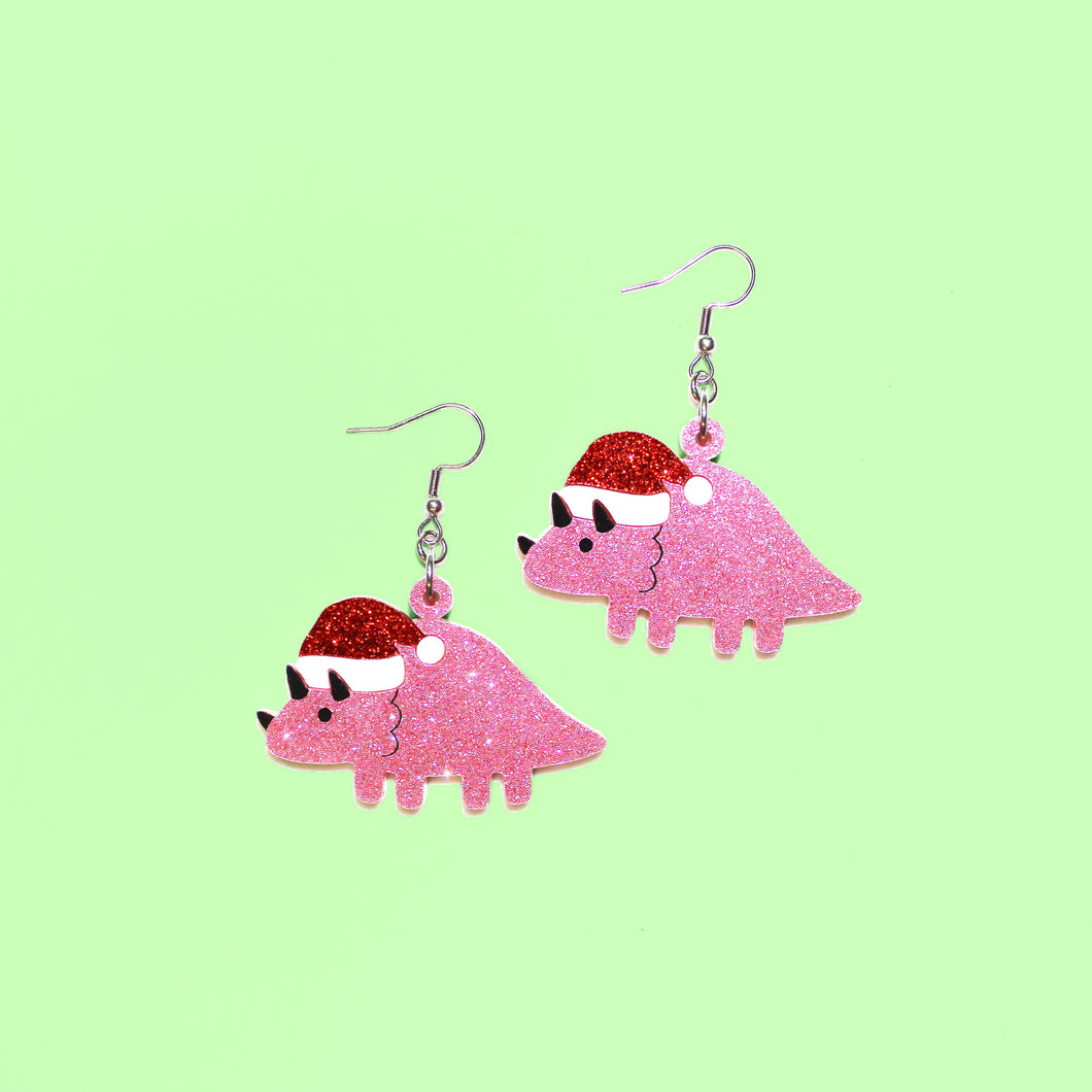 A pair of sparkly dinosaur earrings. The dinosaur is Triceratops, a herbivore that walked on all fours with three distinctive horns on the front of its face and a frill around its neck. This Triceratops is sparkly pink and wearing a sparkly red santa hat. The earrings are made from acrylic, a type of plastic, and hang from silvery coloured stainless steel hooks in front of a light green background. The eyes, horns and frill are hand painted details in black.
