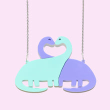 Load image into Gallery viewer, Close up of a dinosaur necklace. The pendant is made of two long necked dinosaurs, like brachiosaurus, one green, one purple, with their necks entwined into sort of a heart shape. They are attached to two silver coloured rings and a fine, stainless steel chain either side.
