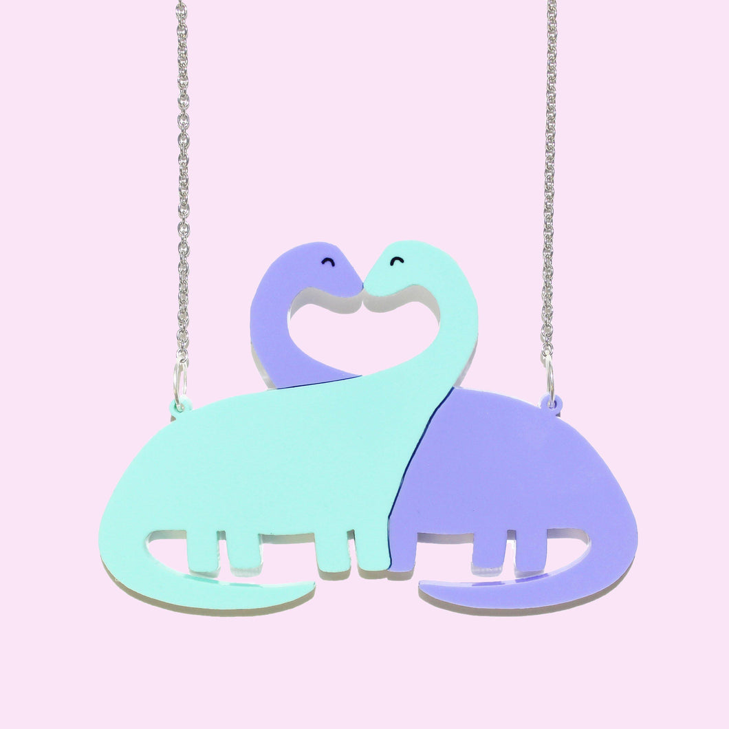 Close up of a dinosaur necklace. The pendant is made of two long necked dinosaurs, like brachiosaurus, one green, one purple, with their necks entwined into sort of a heart shape. They are attached to two silver coloured rings and a fine, stainless steel chain either side.