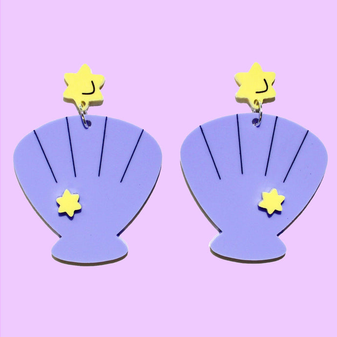 A pair of blueish purple shell earrings with a pastel purple background. Each earring is made of two parts: a small yellow star on top and a pink scallop shell hanging from a ring below it.