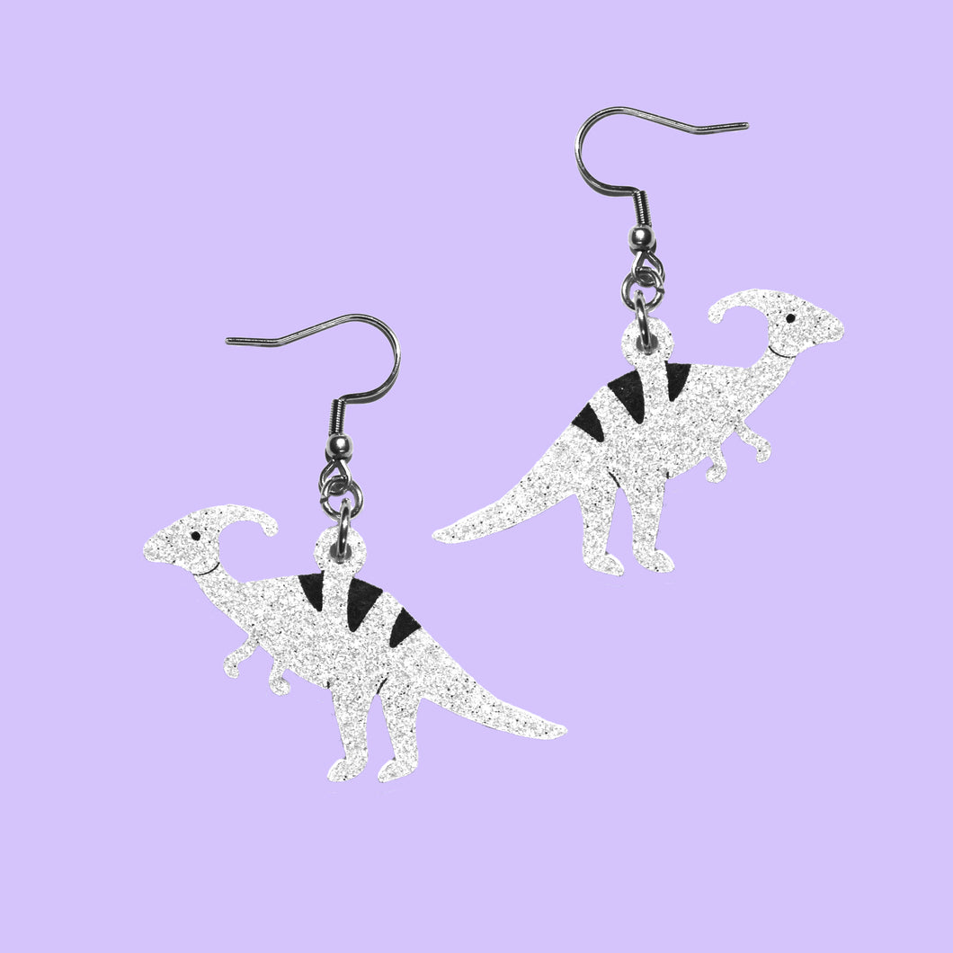 A pair of light silver glitter coloured dinosaur earrings. The earrings are Parasaurolophus, a herbivore that walked around on four legs but could stand on two and has a unique shaped head with a crest on the top that looks a bit like a comma behind it. Think Ducky, from the Land Before Time movie. The earrings are made from acrylic and hang from stainless steel hooks in front of a purple background.
