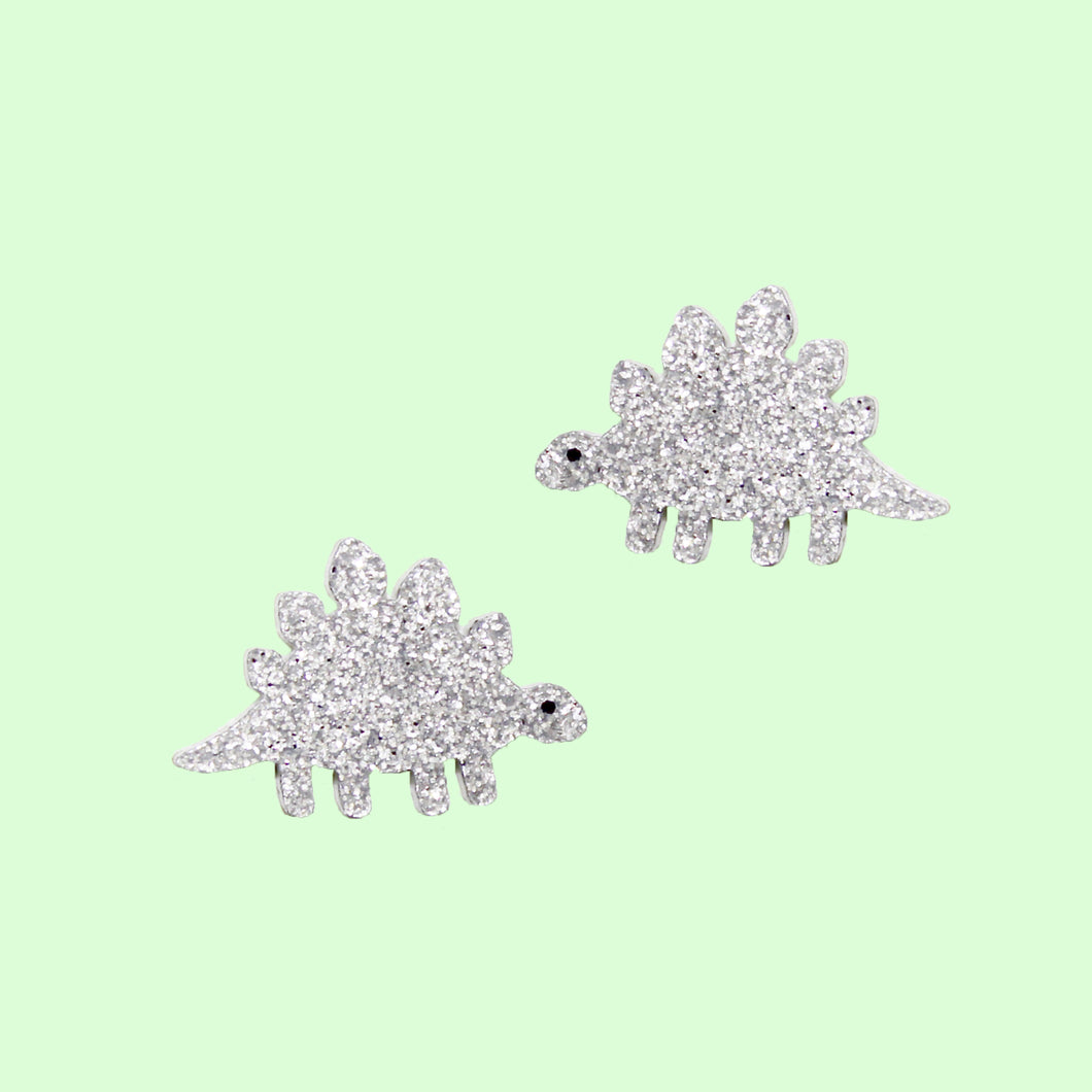 A pair of stud earrings based on the Late Jurassic herbivorous dinosaur, Stegosaurus. The Stego is a light, silvery glitter with hand painted eyes. The stainless steel posts and backs can be seen behind the dino. The earrings are in front of a pastel green background.