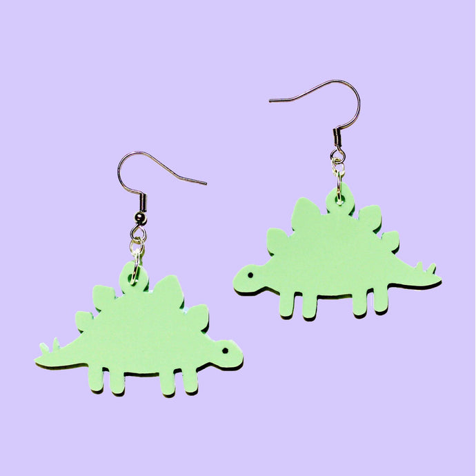 A pair of green stegosaurs earrings in front of a purple background and attached to stainless steel earring hooks.